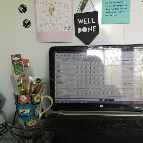 a computer sits on a desk in front of a pin board. The screen shows a detailed spreadsheet, a mug full of pens and hooks sits to the side of the laptop