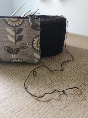 a pencil case holding hooks sits slightly in front of a cake of brown yarn on a wood table, a strand of yarn falls into the foreground