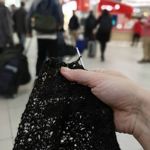 a hand holds a partially completed dark brown lace garment against a blurred background of an airport