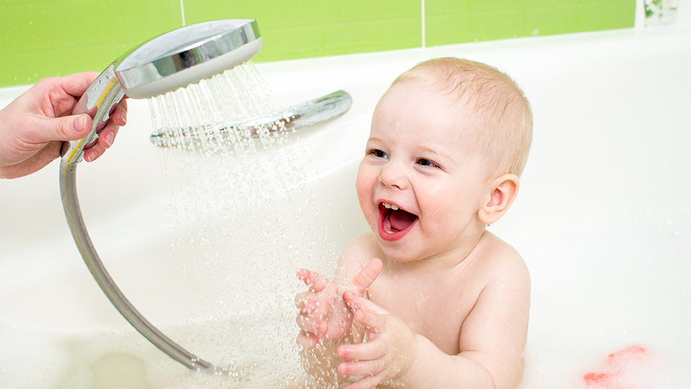 a woman's hand holding a shower head and bathing a smiling baby