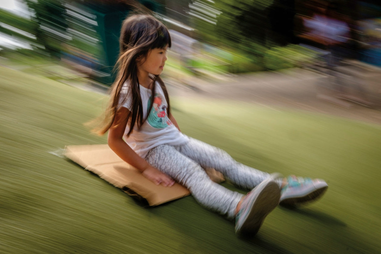A young child slides down a hill, sitting on a piece of cardboard as they fly down the grassy hill. Photo: Jay Tayag
