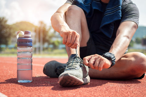 A runner sitting on a red track field tying the laces of athletic shoes with a water bottle in the foreground and a clear sky in the background.