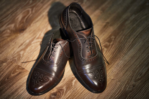 A pair of polished brown leather dress shoes with intricate brogue detailing and thin, dark laces, presented on a wooden floor.