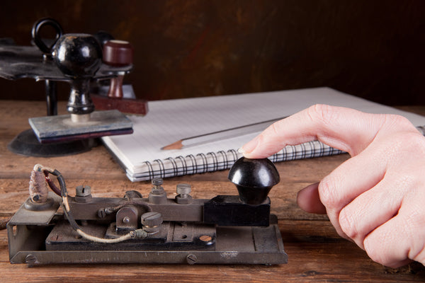 A close-up photo of a hand operating an old-fashioned Morse code telegraph key with a notebook, pencil, and ink stamps in the background on a wooden surface.