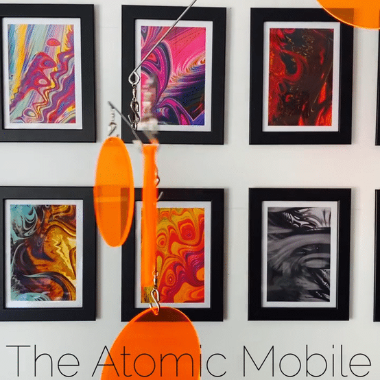 The Atomic Mobile in Clear Orange moving in front of framed colorful abstract art prints - hanging art mobiles by AtomicMobiles.com
