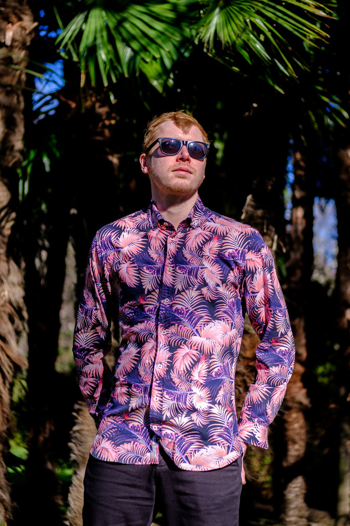 A promotional image of a shirt with a pink and rose palm print featuring hidden tigers. The shirt is made from high quality 100% cotton fabric and is shown hanging on a hanger against a white background. The text reads "Introducing our newest shirt - a stunning pink and rose palm print with a surprise detail.