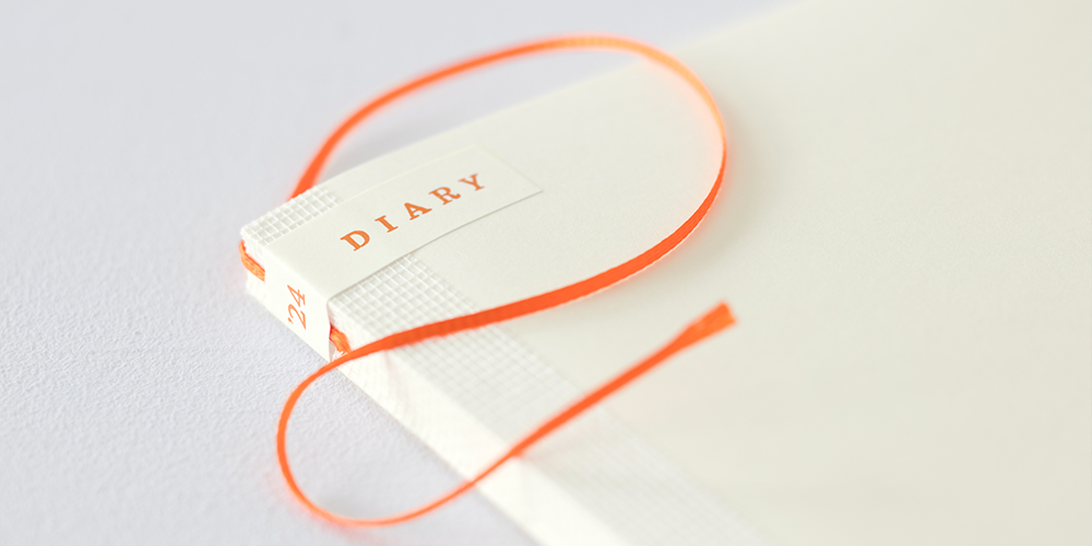 The top left corner of a Midori MD 2024 Diary on a flat grey surface with an orange bookmark draped across the front cover. The words "'24" appears on the spine and the "Diary" appears on the cover.