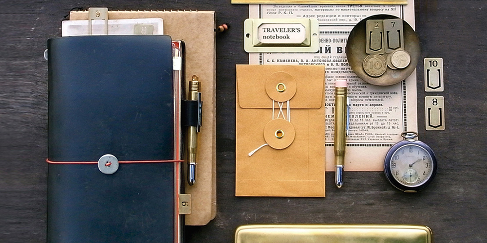 A flatlay of a Blue Traveler's Notebook Regular with two brass traveler's pens and a variety of brass Traveler's accessories on a wooden background