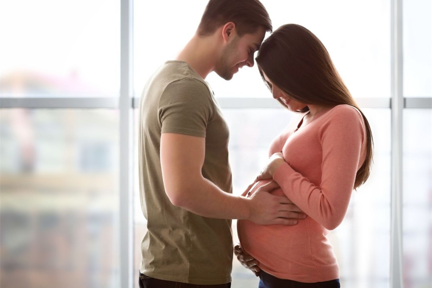 couple maternity photo of  standing and holding each other by the window