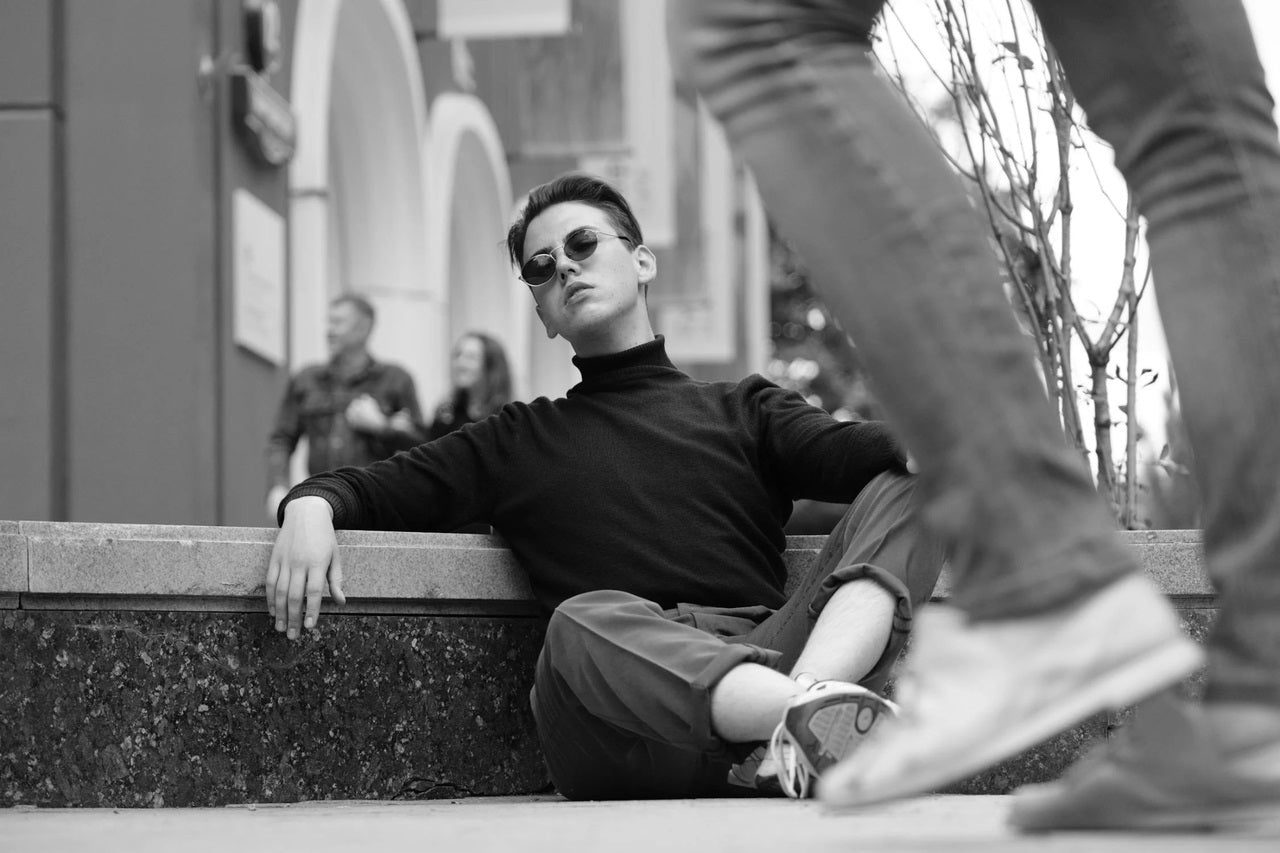 A handsome male model wearing sunglasses is sitting on the street with a cold expression