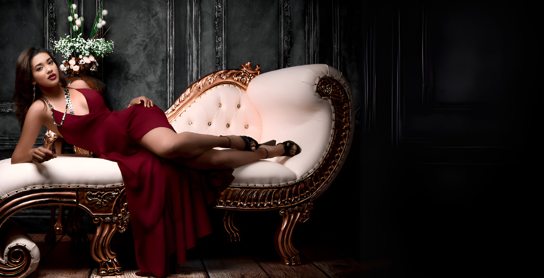  Woman in a burgundy dress reclining on a vintage white and gold chaise against a dark elegant backdrop.
     