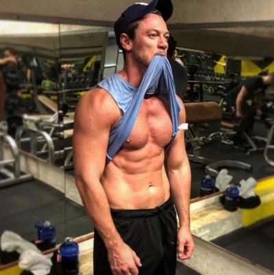 Luke Evans on instagram in the gym black cap and muscles blue vest