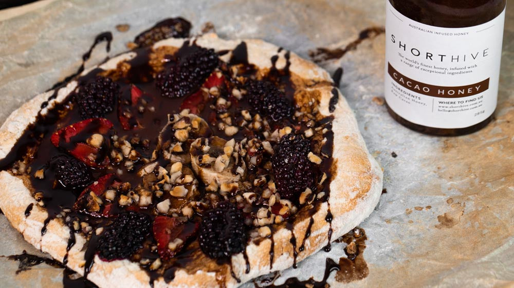 Image of ShortHive Cacao pizza recipe. Fruit chocolate honey drizzled pizza on baking paper with ShortHive Cacao honey bottle on the right