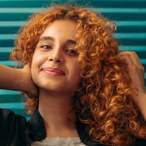 Woman with curly copper hair touching her hair and smiling