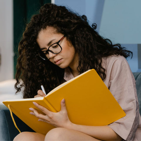 Woman with curly hair and glasses journaling onto a yellow notebook.