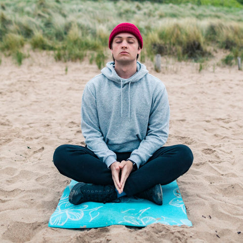 Man sitting at the beach with a beanie on, eyes closed and meditating.