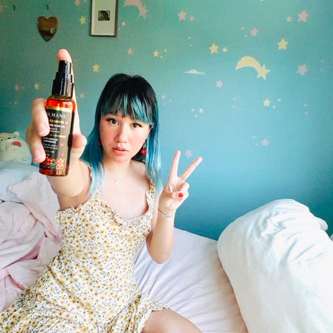 Woman with blue hair holding up a peace sign in one hand and Loba Mane's Illuminating Hair Oil in the other hand.