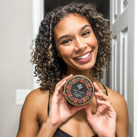 Woman with defined curly hair, holding Loba Mane's Transformative Hair Mask and smiling.