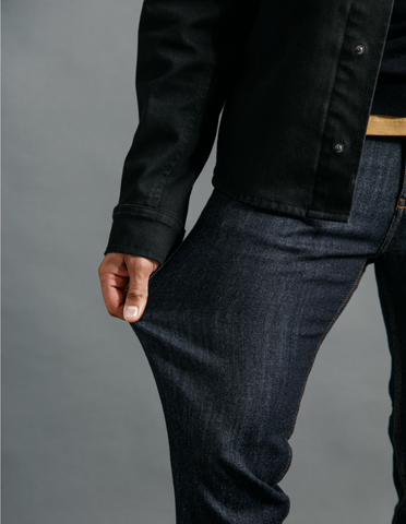 A person wearing a black jean jacket stretching their DUER performance denim