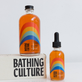 Orange oil in glass jars. One is large and on a platform and another is small and not on a platform. Bottles read: Bathing Culture with a swirly rainbow.