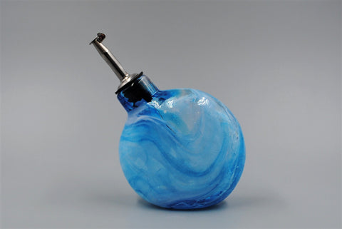 A blue colored hand blown oil bottle with a stainless steel spout, shown facing the left. 