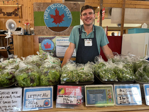 Jordan MacPhee, owner of Maple Bloom Farm, standing behind our booth at the Charlottetown Farmers’ Market in front of fresh lettuce and veggies