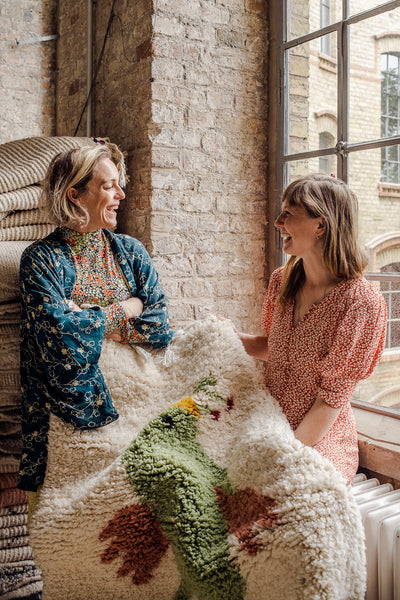 Two women sitting by a loft window holding a berber rug with a bird design.