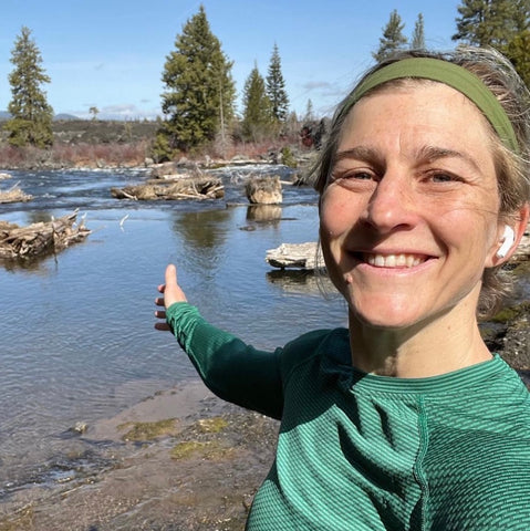 Lauren Fleshman standing in front of a shallow river, pine trees in the distance, wearing a green long sleeve top and head phones, smiling with a hand outstretched towards the water