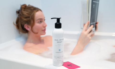 Woman reading a book in a bubble bath with a bottle of Graydon Skincare All Over Soap on tub ledge