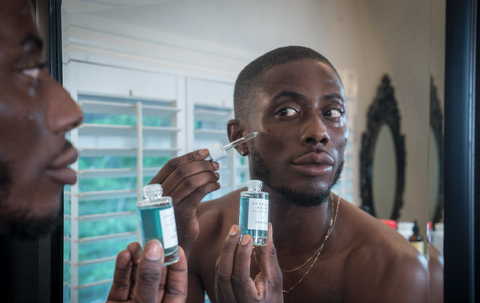Man looking in mirror, holding a bottle of bright blue Graydon Skincare Fullmoon Serum and applying product to face using dropper.