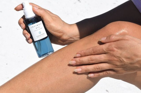 Close up of woman holding bottle of bright blue Graydon Skincare Intimacy Oil over leg and rubbing oil into skin.