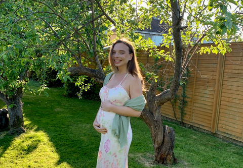 Pregnant woman in floral dress, standing on the grass, holding belly.