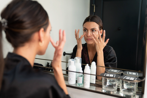 Brunette woman looking in the mirror and applying eye cream using her ring fingers.