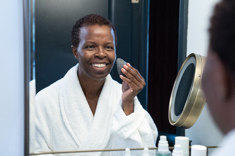 A dark-skinned woman in a comfortable white robe cleansing her skin in front of a mirror using a bamboo charcoal sponge