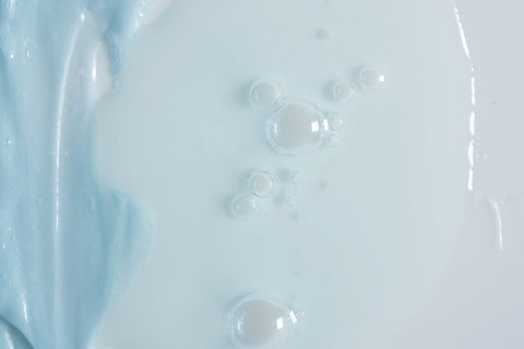 A smear of blue-hued probiotic face cream next to several drops of an aquamarine-coloured retinol alternative face serum on an off-white surface