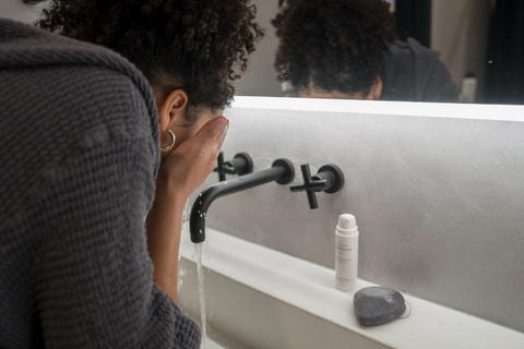 Woman washing her face. Leaning over a sink with running water and a bottle of Graydon Skincare Aloe Milk Cleanser and a Bamboo Charcoal Sponge next to the faucet.