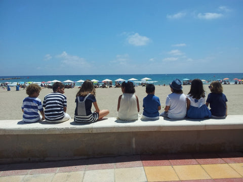The image is of eight young children sitting on a concrete ledge overlooking the beach. The children appear to be talking to each other while the beach is relatively busier closer to the water, but the children are seated quite a distance from the water.