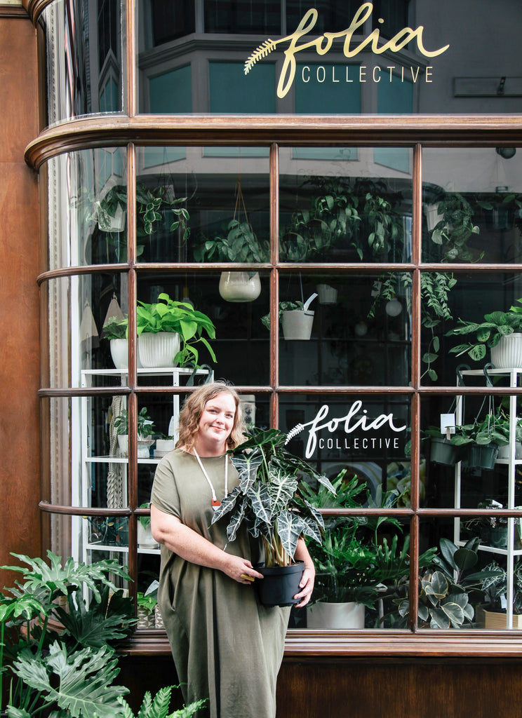 Danae, the owner of Folia stands outside the first Folia shop, holding a plant. She is wearing a green dress, and has shoulder length blonde hair.