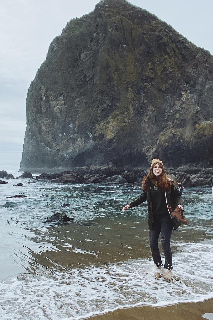 A woman with red hair stands in shallow ocean surf, with a very large rock rising from the water in the background