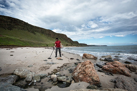 image of man metal detecting on a beach with hillside in background