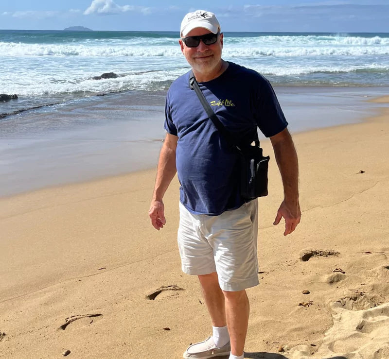 Man in casual attire standing on a sandy beach by the ocean.