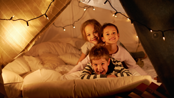 Three children smiling under a blanket fort with pillows and string lights.