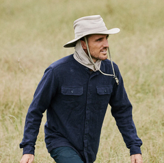 Man wearing a sailing hat with a neck flap and a blue long-sleeve shirt walks through a field of grass