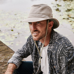 Man wearing a recycled plastic fedora and printed button down sits next to a pond