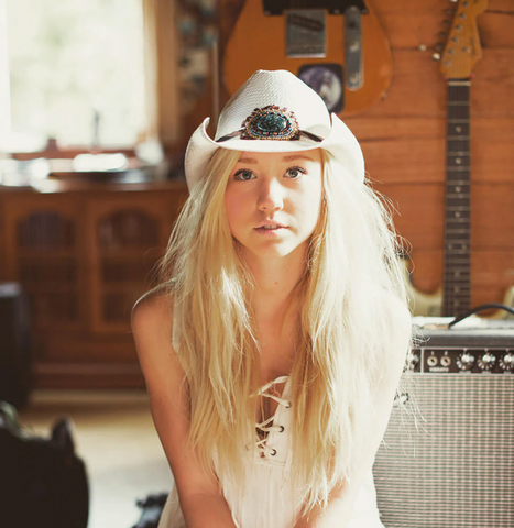 blonde woman wearing a white western hat and a white dress stares into the camera