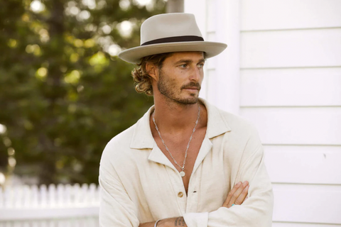 Man wearing a tan wool fedora and a white button down shirt stares to the left with his arms crossed