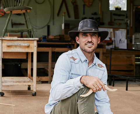 Man wearing a denim button down and leather outback hat kneels on the ground, smiling at the camera. A workshop is in the background.