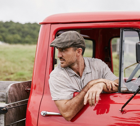 A man sits in a vintage red pickup truck wearing a newsboy cap.