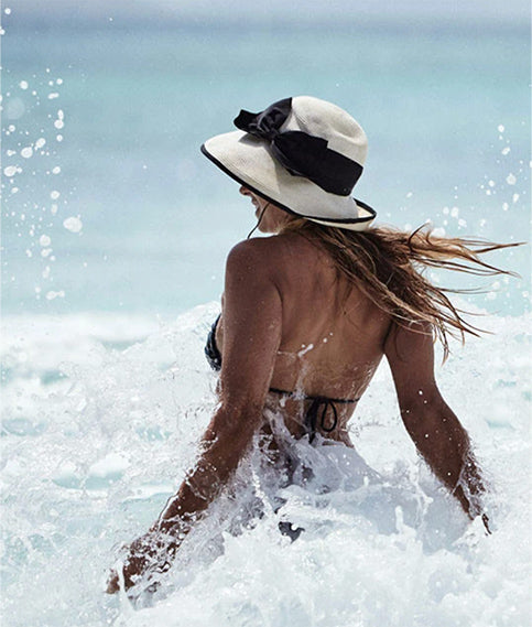 Young woman wearing a Sun Protective hat standing among waves and ocean foam.