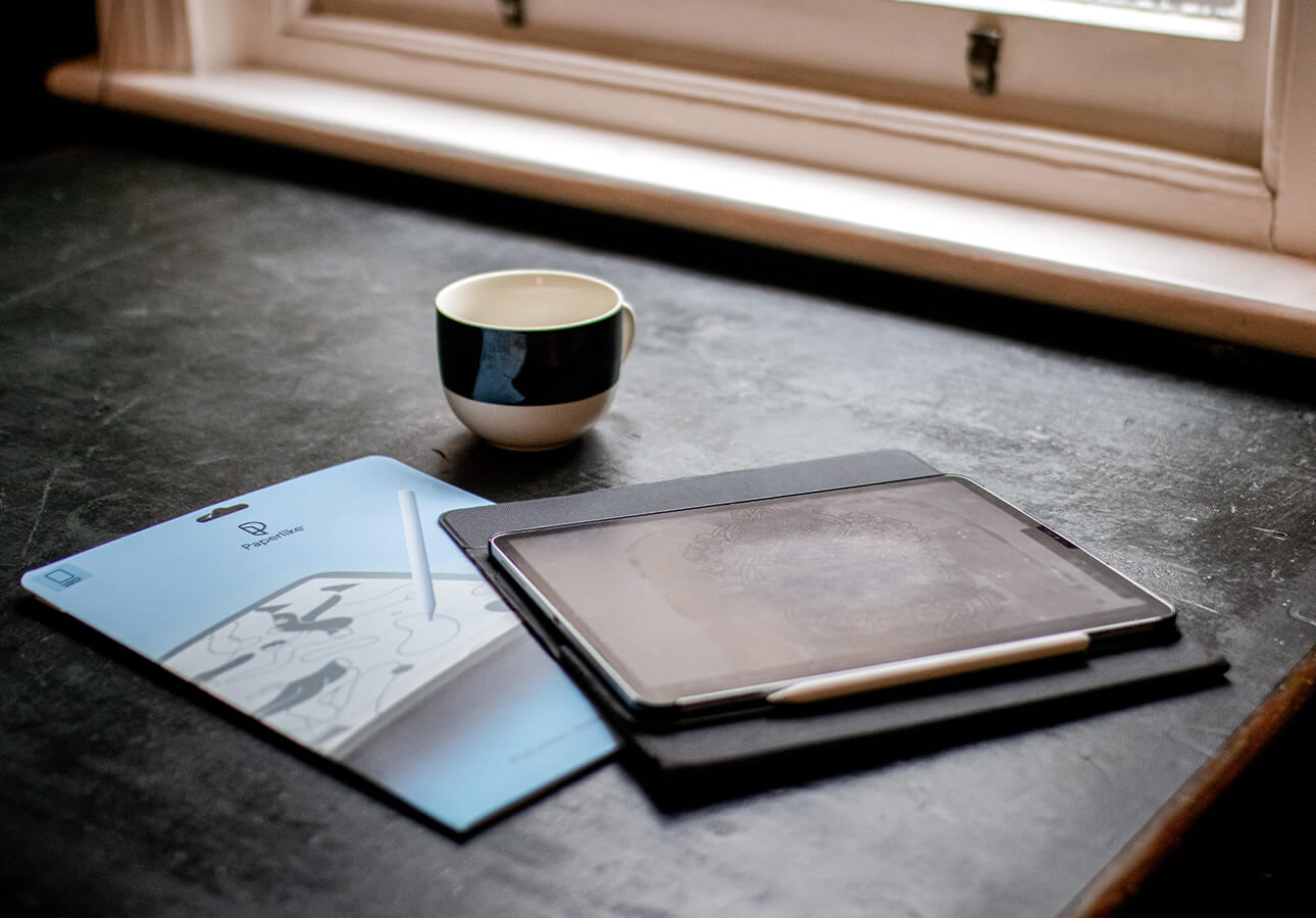 An image of an iPad, Paperlike brand packaging, and a coffee mug situated on a table by a window.